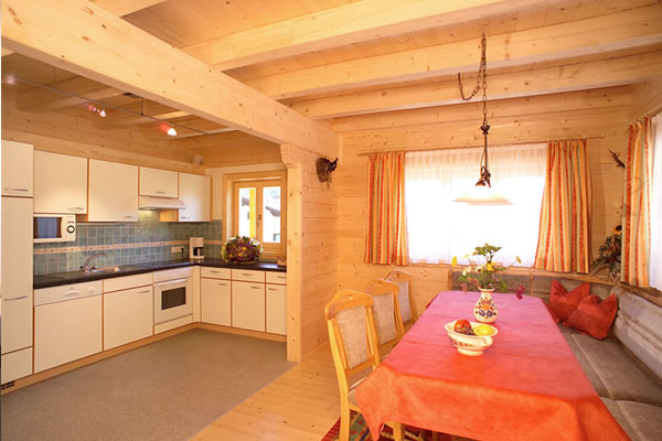 Küche und Essbereich im Chalet 'Jagdhaus' mit Holzbalken, weißen Schränken, blau gefliester Arbeitsfläche, Esstisch mit rotem Tuch und Fenster mit orangefarbenen Vorhängen. Jagdhaus, Tannenhof Wagrain