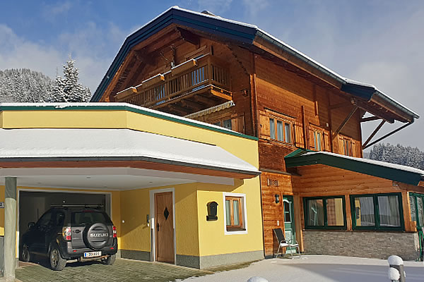 Holzchalet 'Jagdhaus Wagrain' mit gelbem Anbau, Balkon und schneebedecktem Dach, davor schwarzer Suzuki-Pkw unter Carport.