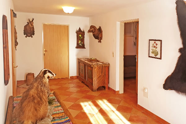 Flur im Chalet 'Jagdhaus Wagrain' mit braunem Fliesenboden, Holztür, antiker Kommode, Wanduhr und Tierpräparaten an den Wänden.