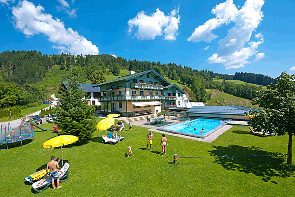 Ferienwohnungen Tannenhof in Wagrain mit grünem Garten, Schwimmbad, Sonnenschirmen und Menschen bei sonnigem Wetter vor bewaldeten Hügeln. Aktiv im Sommer - Ferienwohnungen Wagrain