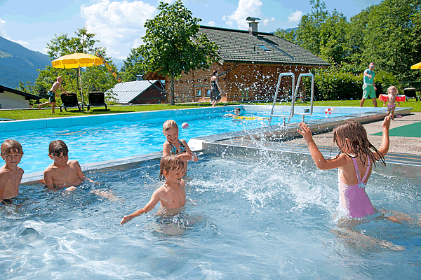 Kinder spielen im Außenpool der Ferienwohnungen Tannenhof in Wagrain, umgeben von grünen Bäumen und Bergen im Hintergrund.
