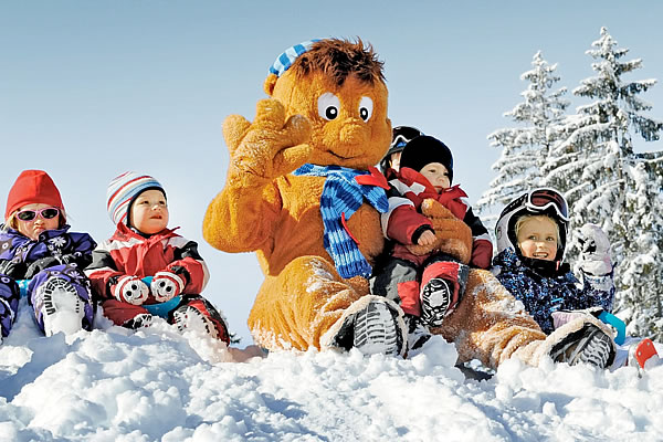 Große braune Stofffigur der 'wagraini' mit blauer Mütze und Schal sitzt im Schnee, umgeben von Kindern in bunter Winterkleidung und Skibrillen, Tannenbäume im Hintergrund.